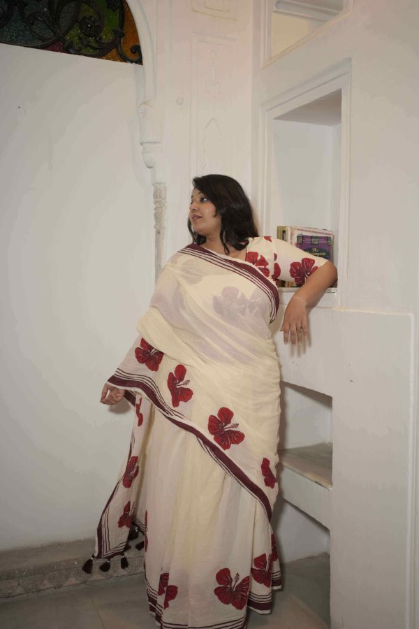 Surmayi cotton saree in cream with red hibiscus motifs along the hem, maroon striped border, tassels, and matching hibiscus print blouse fabric.
